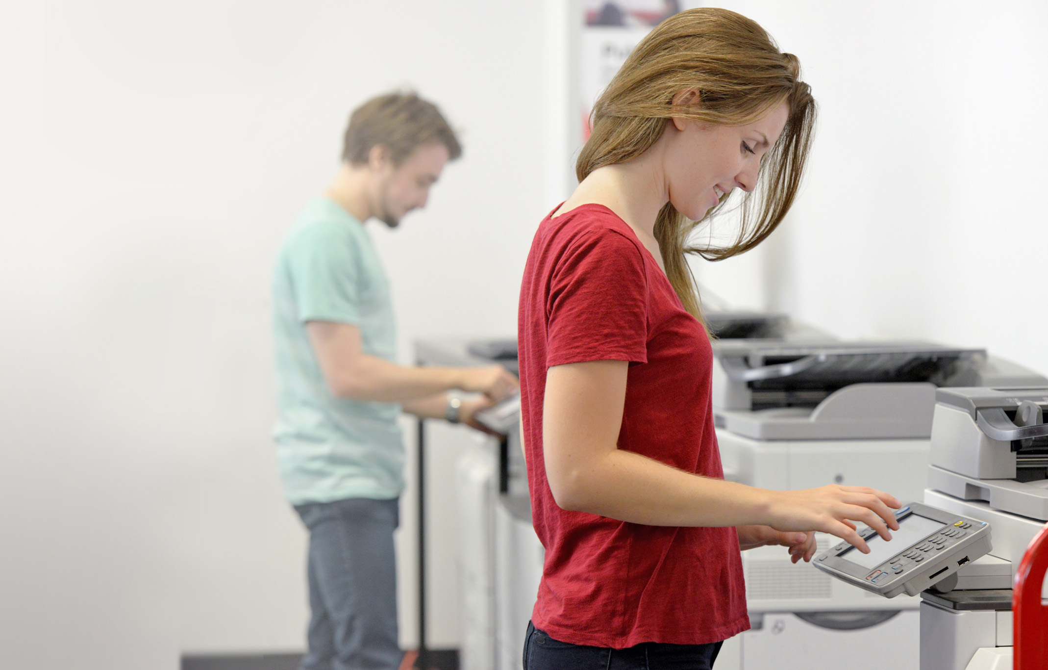 Woman in red shirt at fax machine with man in light green shirt in the background at another fax machine